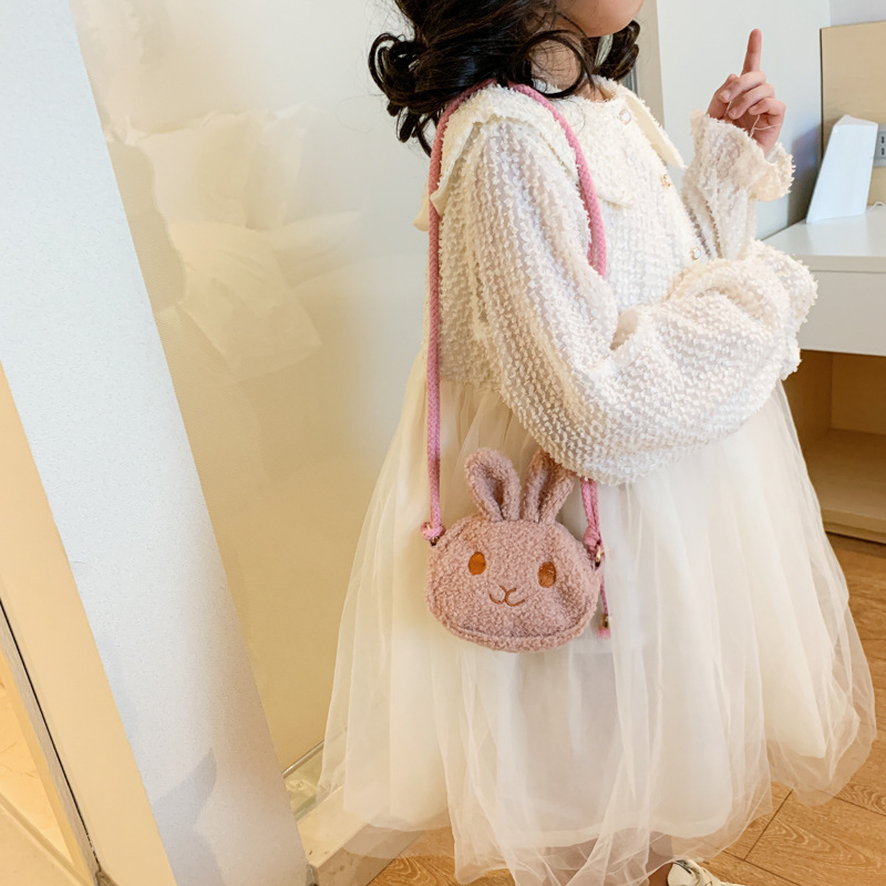 Petite fille de profil, qui porte une robe blanche et un sac en peluche en tête de lapin beige qu'elle porte sur son épaule