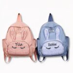 2 petits sacs à dos personnalisés aux prénoms de Ylla et Kathlee. L4un est bleu, l'autre rose et ils ont un motif d'oreilles de lapin devant, sur fond gris clair