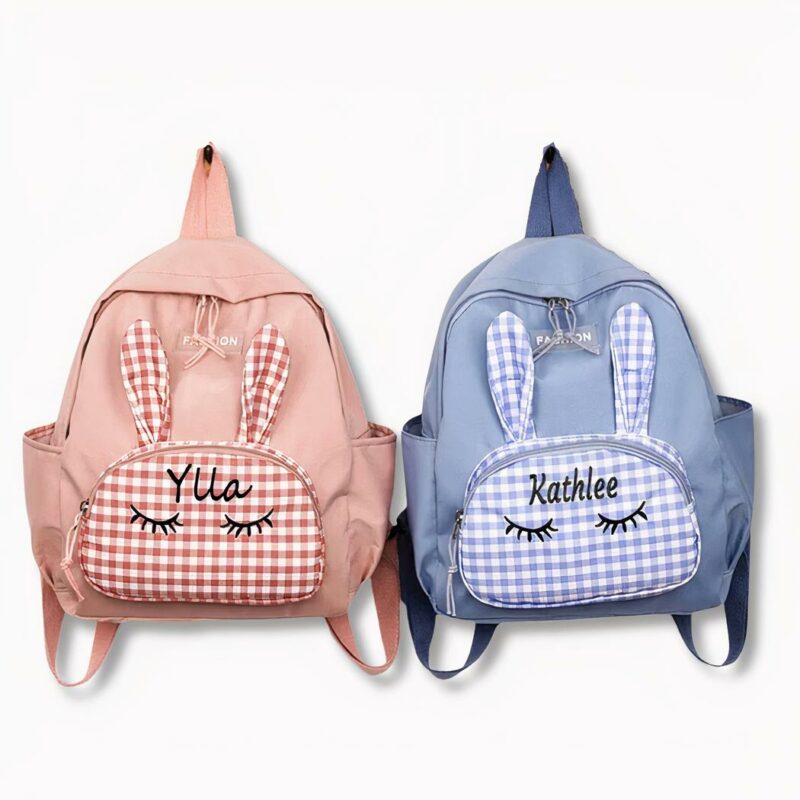2 petits sacs à dos personnalisés aux prénoms de Ylla et Kathlee. L4un est bleu, l'autre rose et ils ont un motif d'oreilles de lapin devant, sur fond gris clair