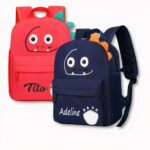 2 sacs à dos pour enfants, l'un rouge et l'autre bleu foncé avec des visages de dinosaures souriants et personnalisés au nom de Tito et Adeline, sur fond gris clair