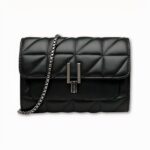 Sacoche à bandoulière pour femme. Elle est noire avec un style matelassé en similicuir. Elle se ferme avec un fermoir en métal noir, et se porte grâce à une sangle en chaîne. Le fond de l'image est gris clair.