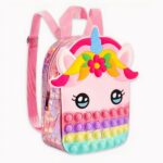 Photo d'un sac à dos rose à tête de licorne. Le contour du sac est brillant, le bas du sac contient des pop it de couleurs rose, violet, jaune et bleu. Au milieu, il y a ses yeux et au-dessus ses oreilles avec une crinière en forme de fruits et fleur, puis sa corne qui est bleue. Le fond de l'image est gris clair.