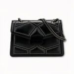 Sacoche à bandoulière pour femme, ayant comme design un style perlé géométrique. La sacoche est noire en similicuir, avec une sangle en chaine. La sacoche se ferme avec un rabat. Le fond de l'image est gris clair.